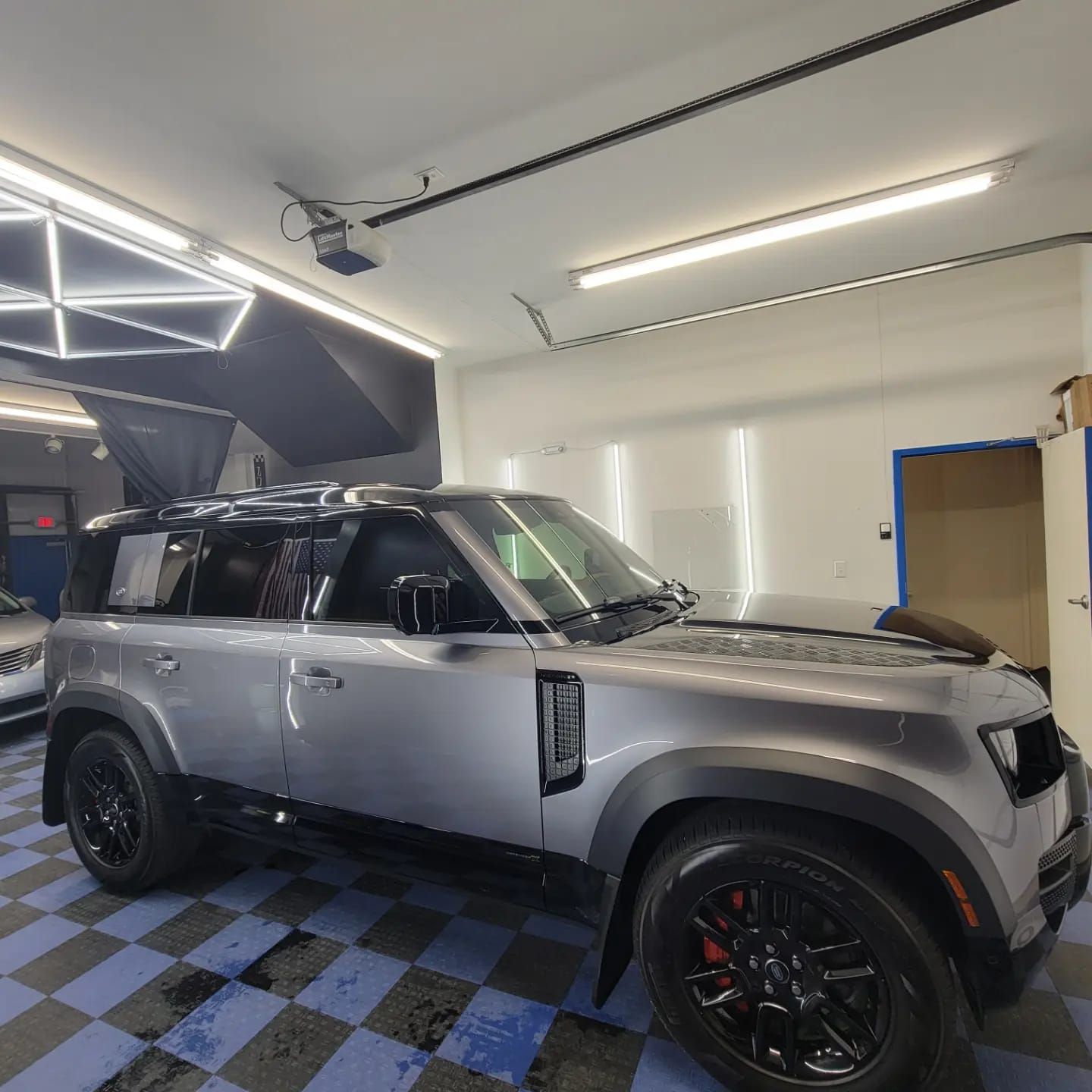 A silver Land Rover Defender SUV parked on a blue and black tiled floor in a brightly lit garage.