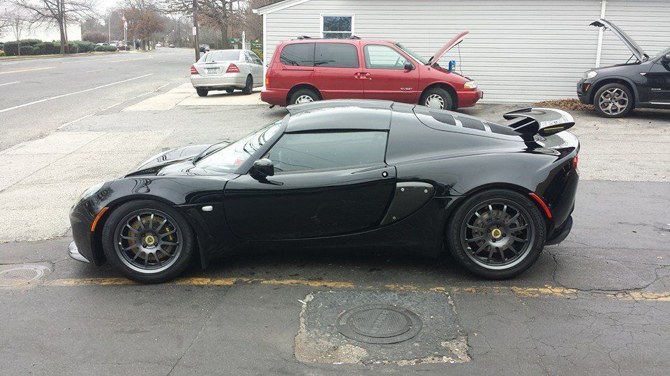 A black Lotus sports car parked in a lot, viewed from the side, with other vehicles in the background.