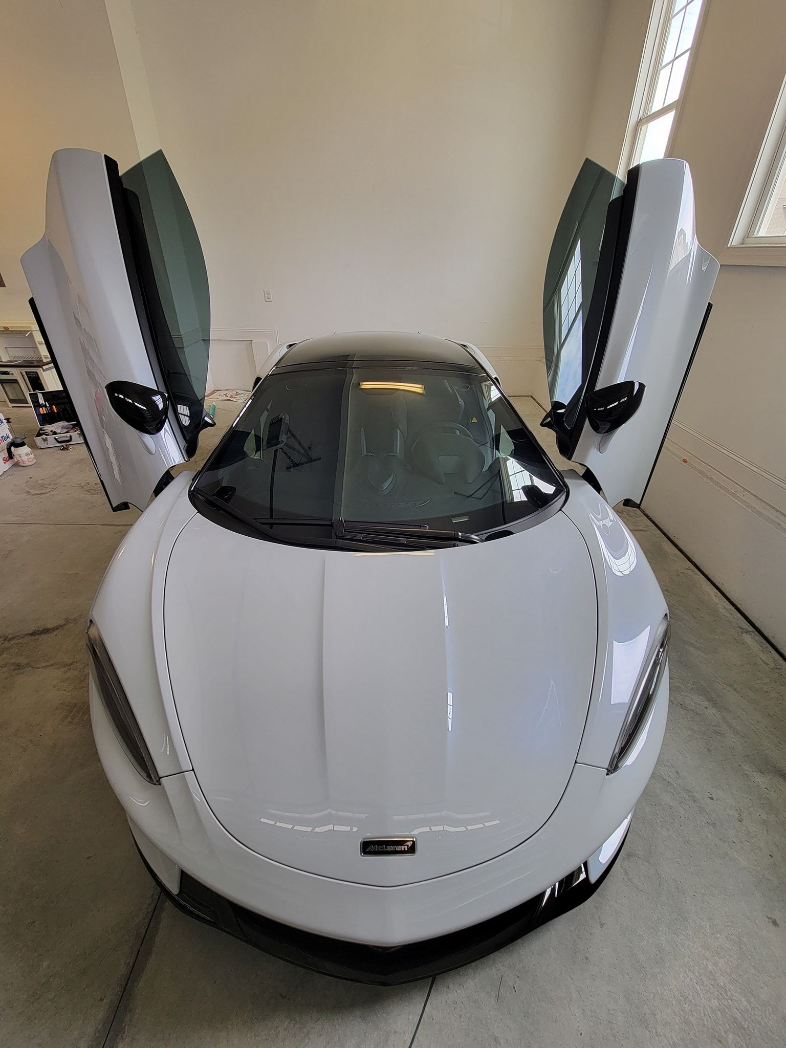 A white McLaren supercar with its dihedral doors open, parked inside a brightly lit indoor space.