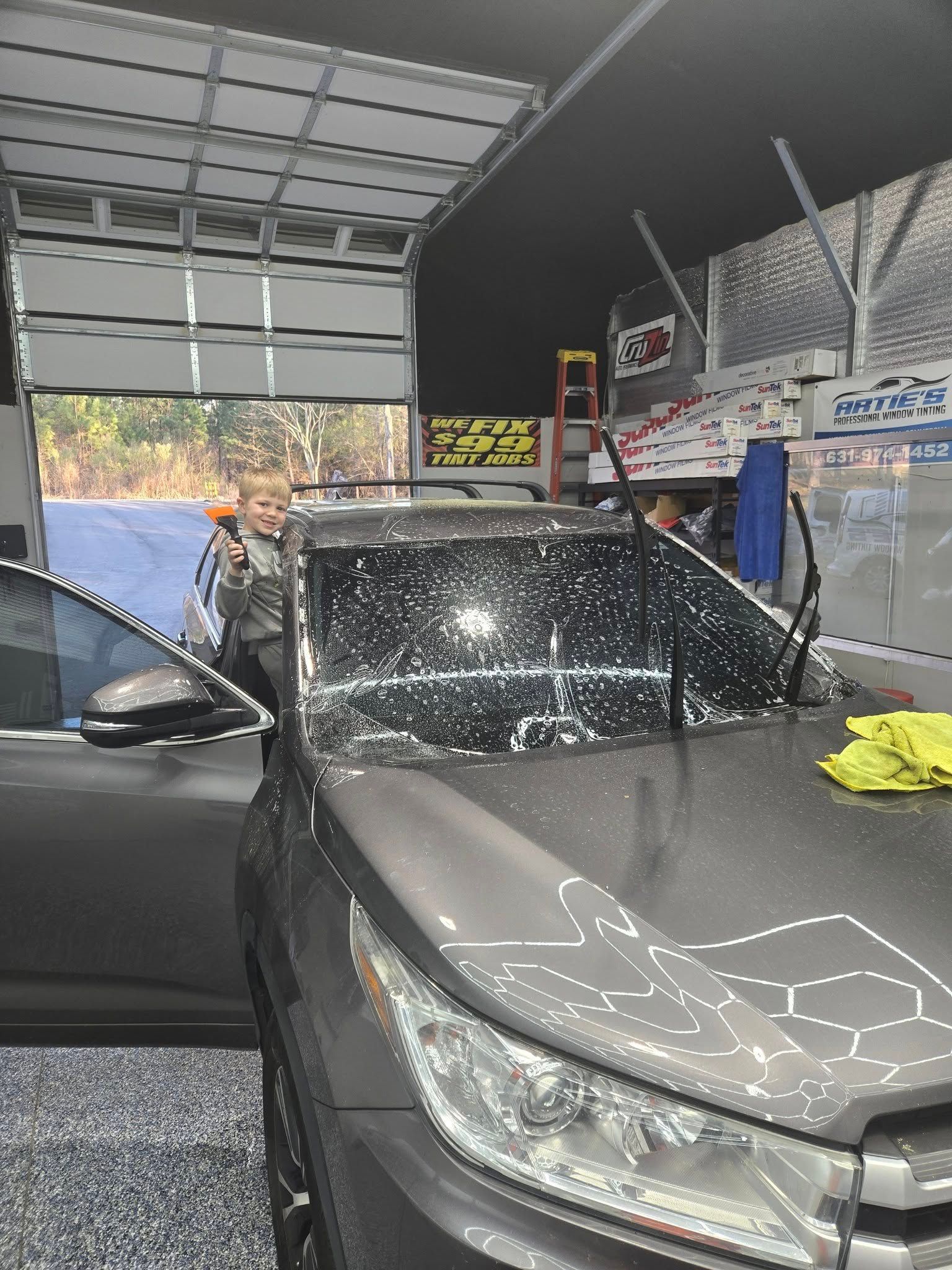 A person works on tinting the windshield of a grey vehicle inside a shop, with the car hood visible in the foreground.