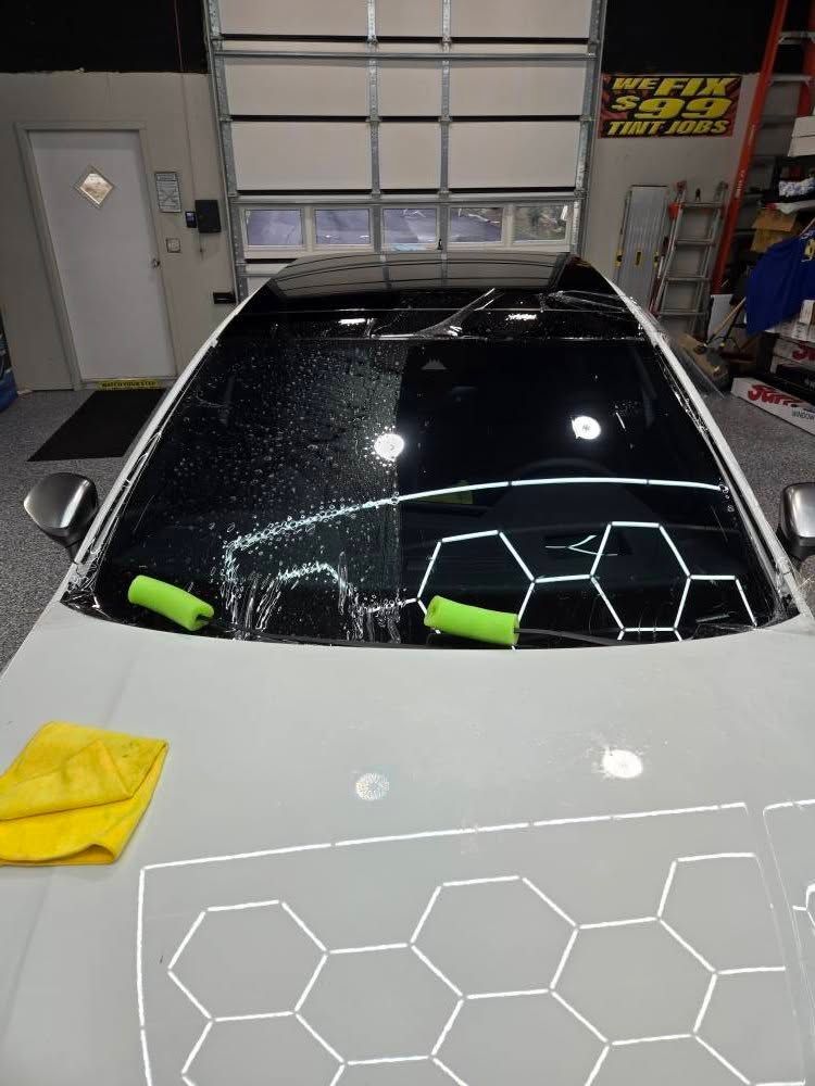 A high-angle shot of a white car’s hood and windshield in a shop, reflecting a hexagonal overhead LED light pattern.