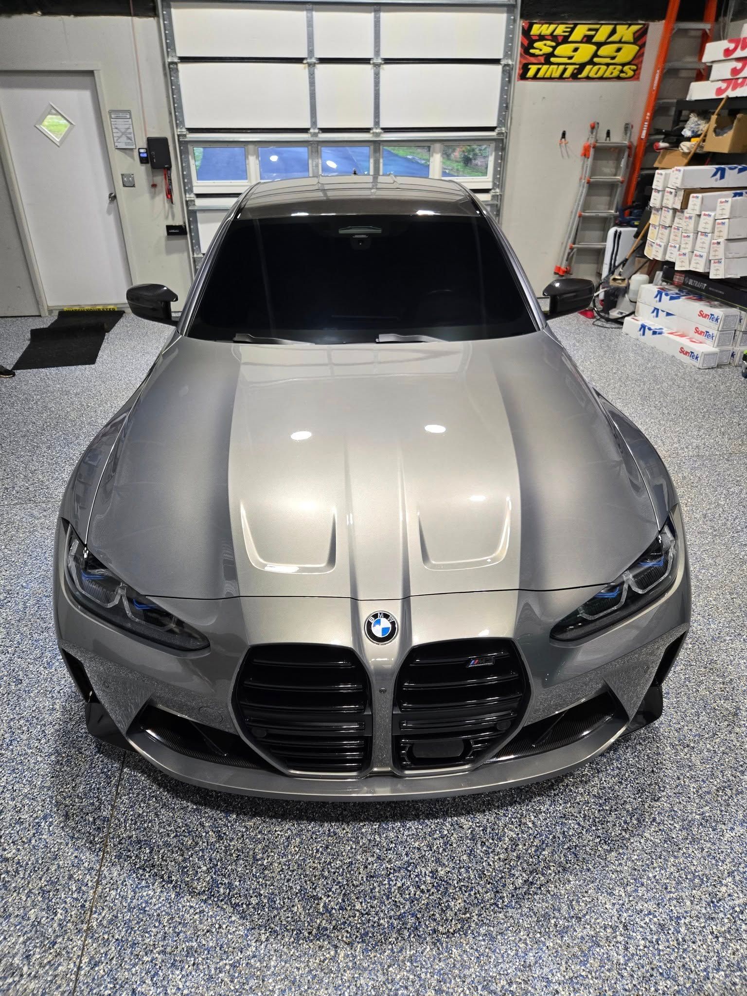 A high-angle view of a grey BMW M4 parked in a garage with a speckled floor and a large, closed roll-up door.