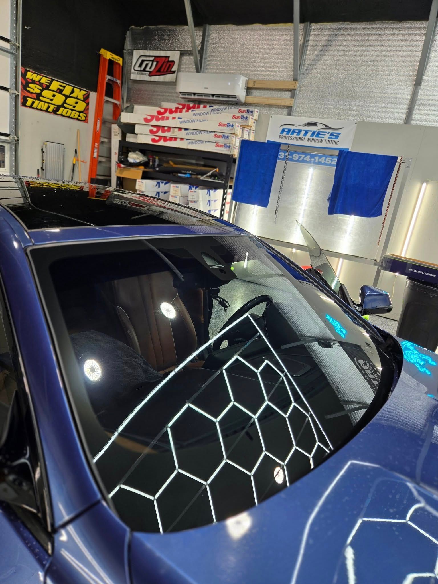 A blue car in a garage with a honeycomb pattern light reflected on its windshield and hood.