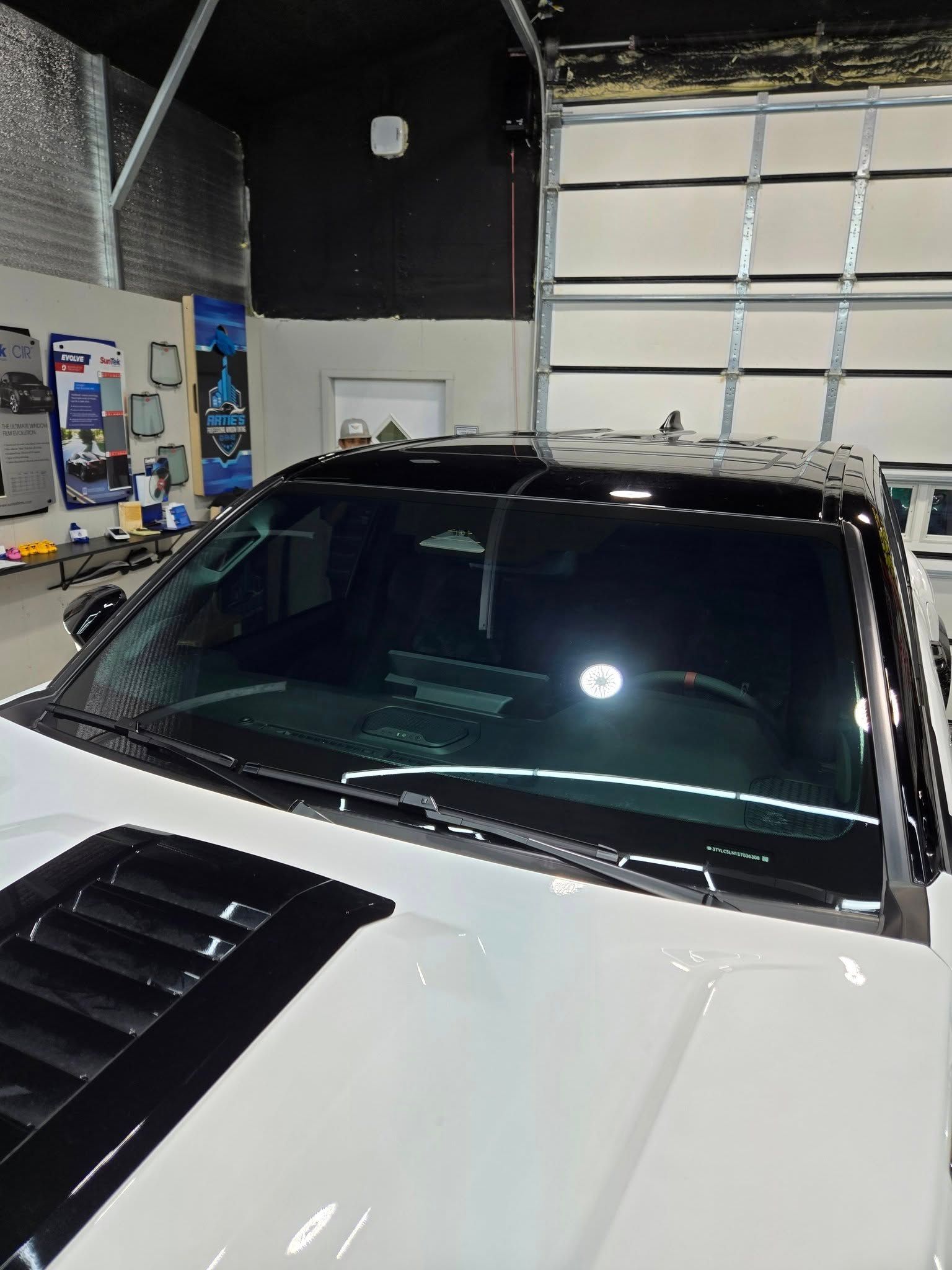 Close-up of a white truck hood and windshield with a black roof and hood vent, parked inside a brightly lit garage.