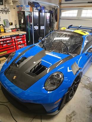 A bright blue Porsche sports car with carbon fiber hood accents parked inside a cluttered garage workshop.