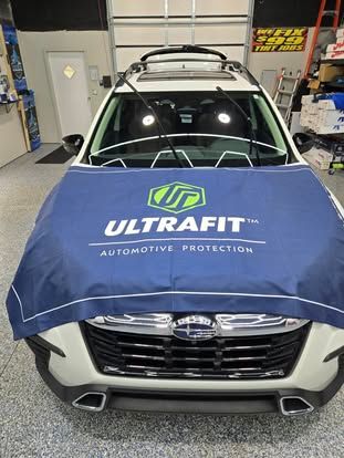 A white Subaru with a blue Ultrafit automotive protection cover over its hood inside a garage.