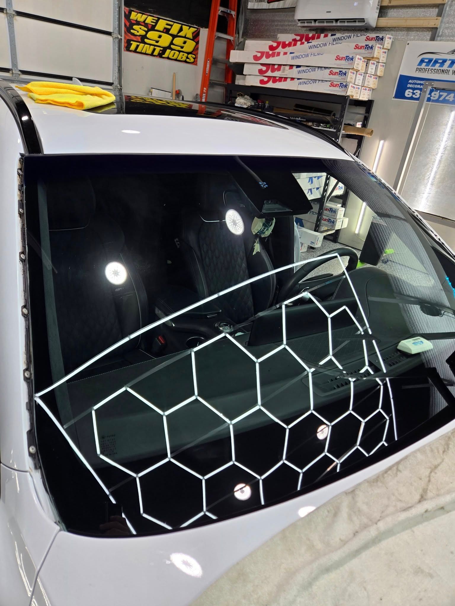 A white hexagonal grid pattern is reflected on the windshield of a car in a workshop, used for assessing glass clarity.