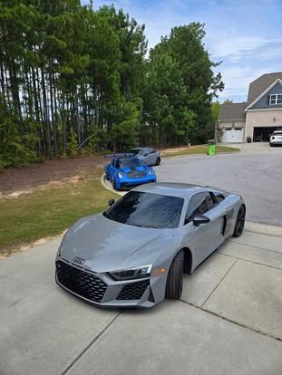 A gray Audi R8 parked on a concrete driveway with a blue sports car parked behind it near a wooded area.
