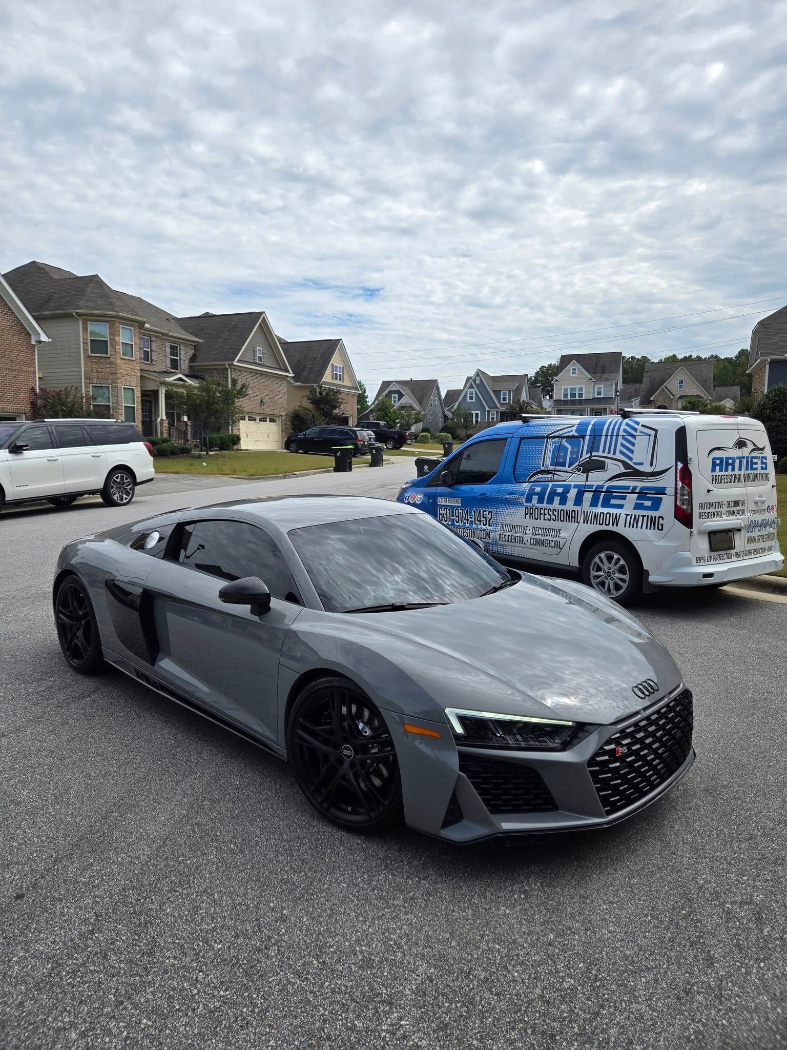A gray Audi R8 parked on a residential street next to a white van with blue logos.