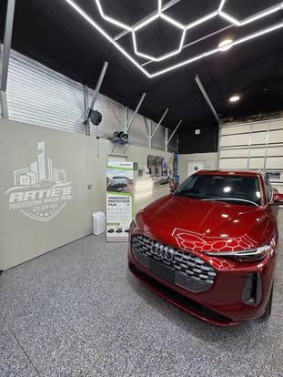 A shiny red Audi parked inside a modern auto shop with honeycomb ceiling lights, a logo on the wall, and a banner stand.