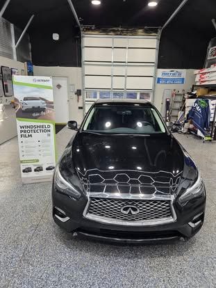 A black Infiniti sedan parked in a garage next to a promotional sign for windshield protection film.