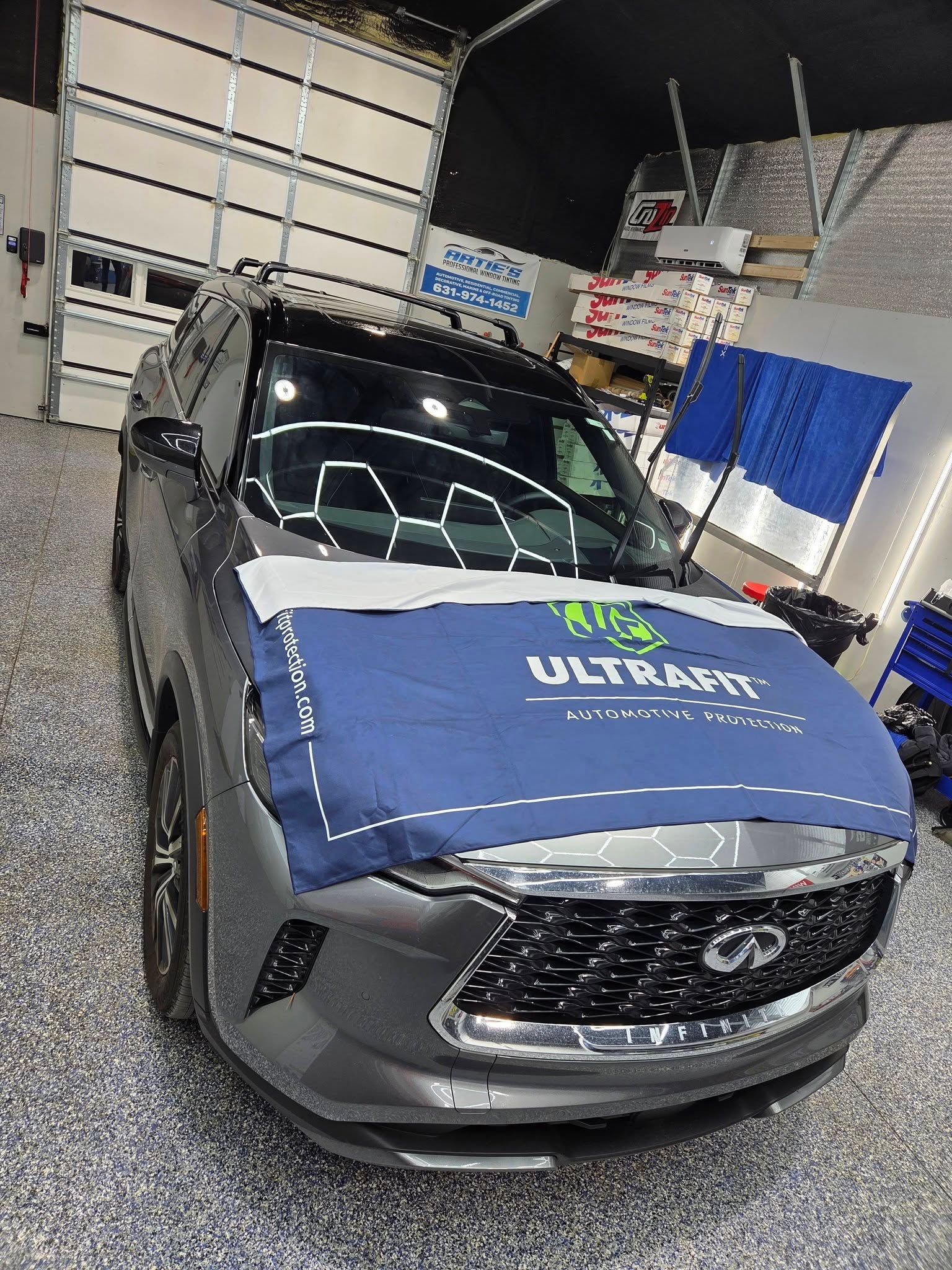 A gray Infiniti SUV in a garage, featuring a blue Ultrafit protective fender cover draped over the hood.