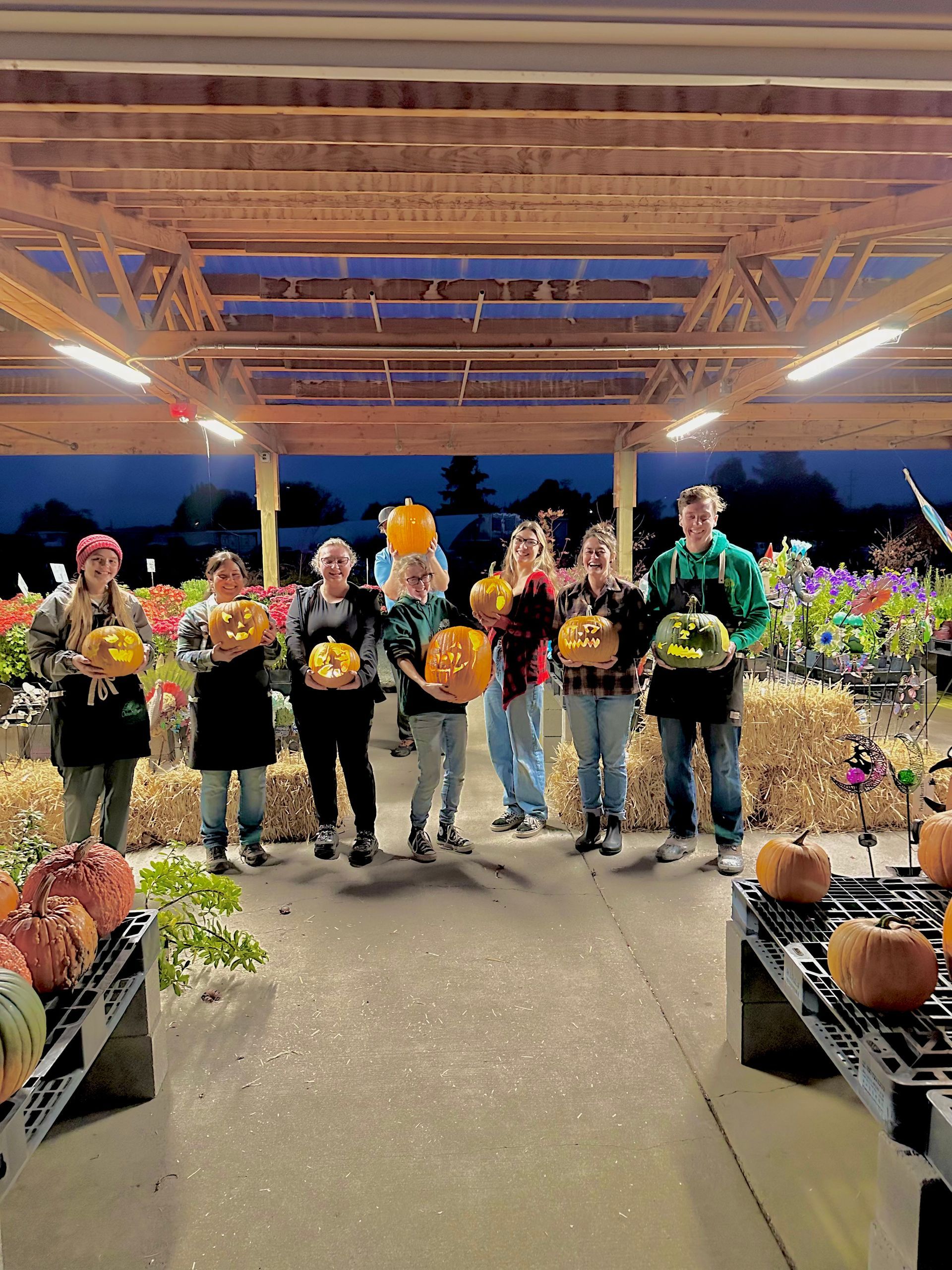 Group of people holding carved pumpkins, posing outdoors under a shelter with lights.