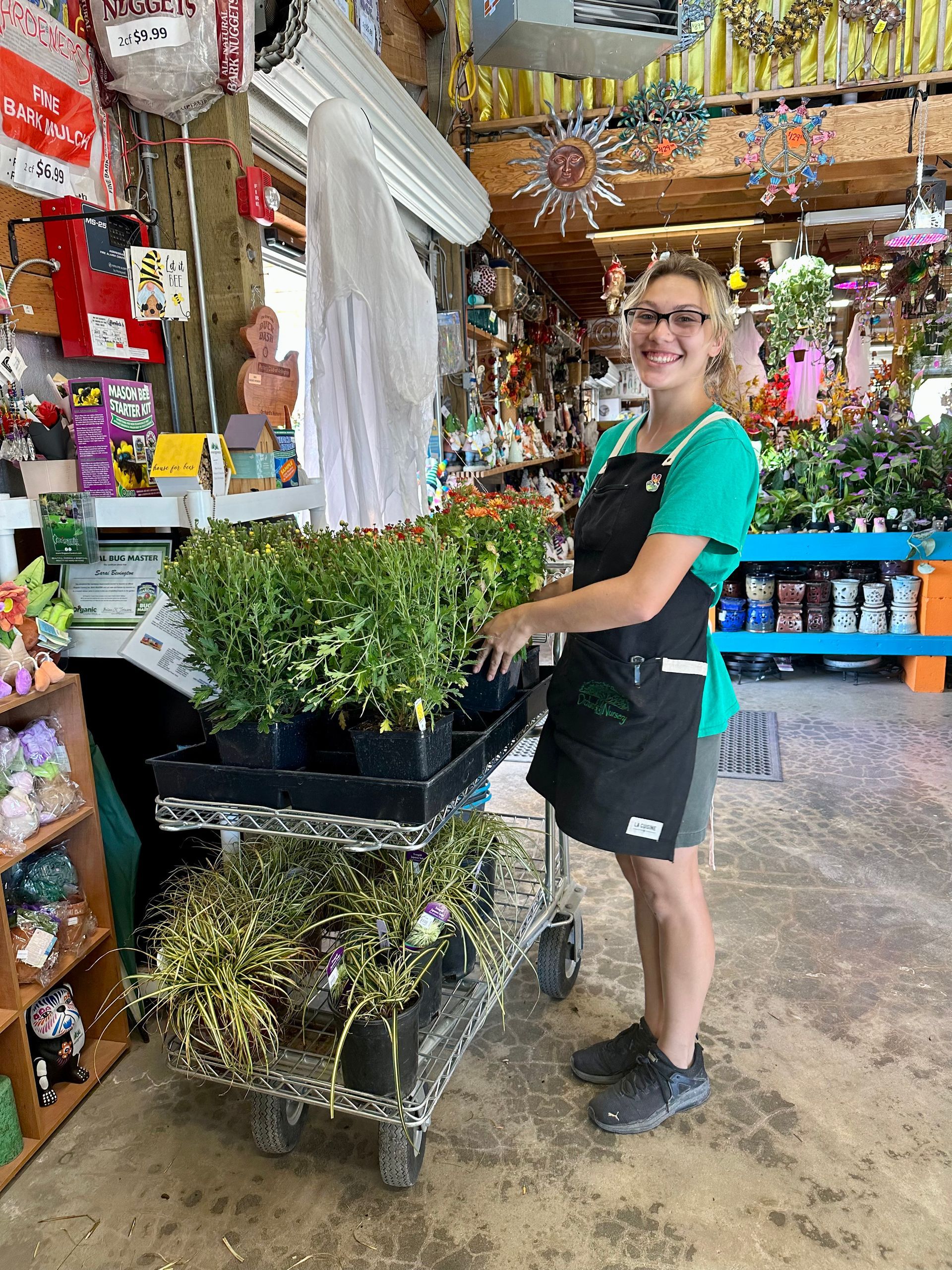 Woman in apron in a plant shop smiles, tending plants on a cart.