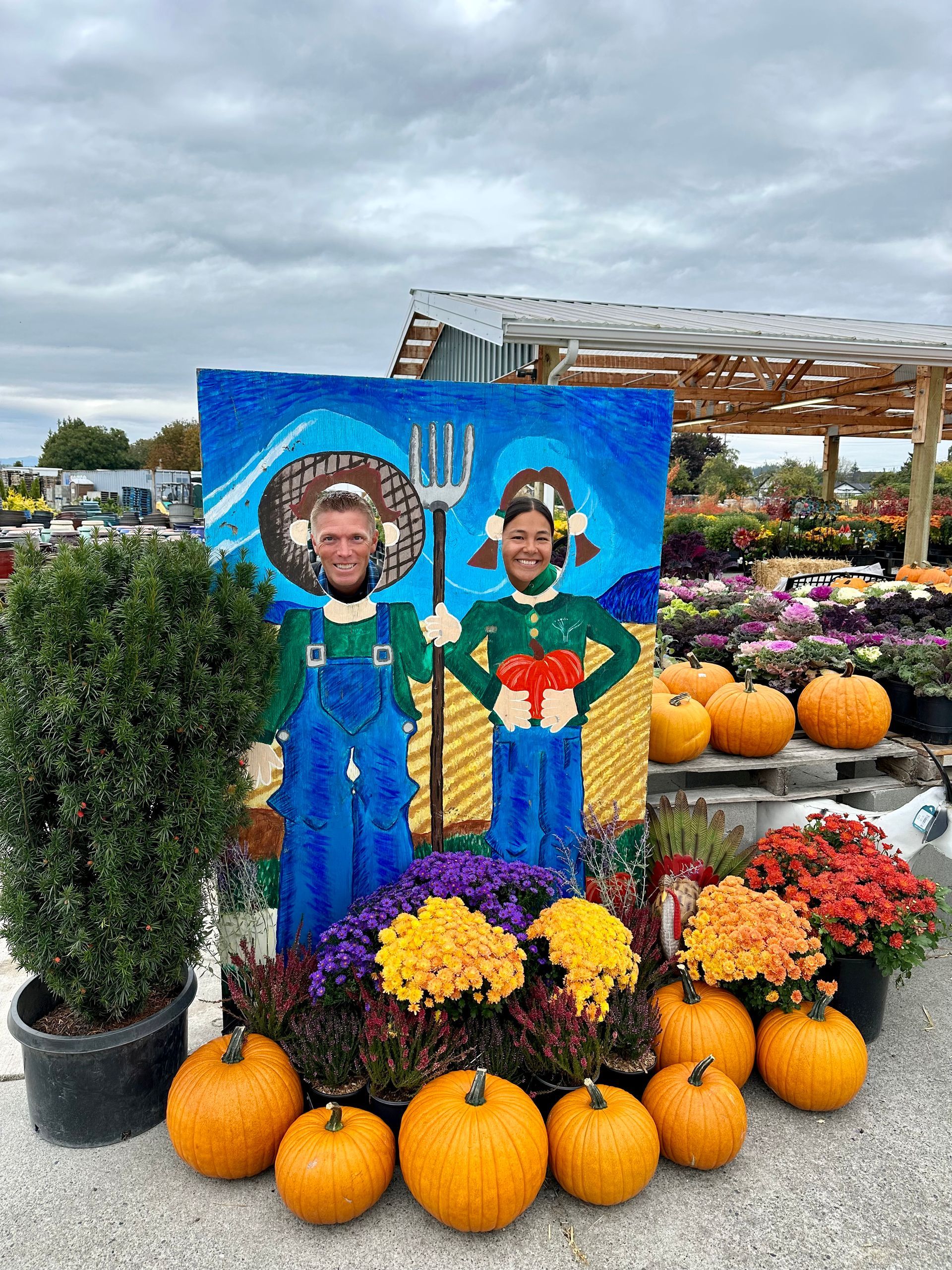 Two people posing in farmer cutout with pumpkins and flowers in front.