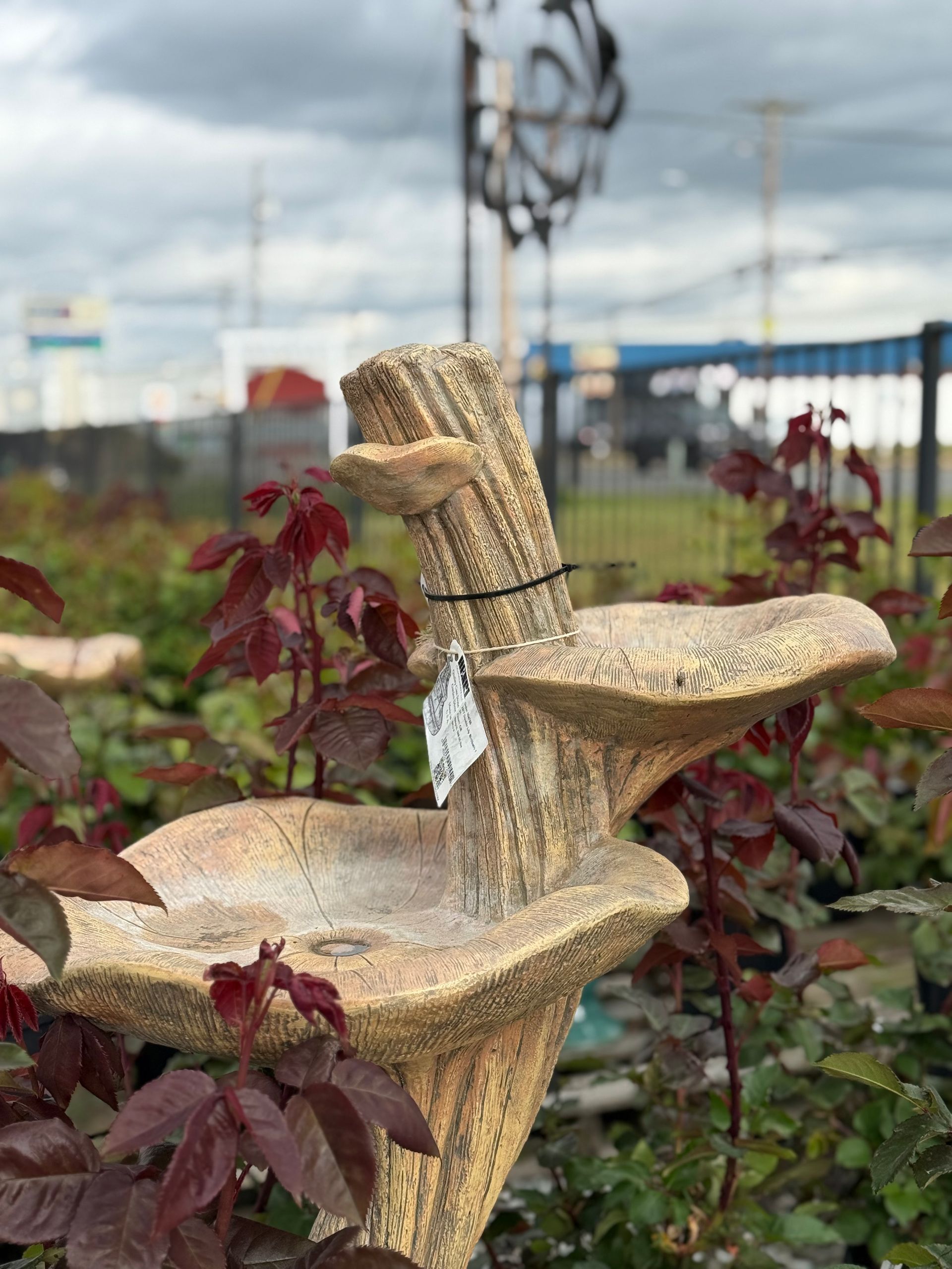 Stone bird bath with tiered bowls, surrounded by plants, against a blurry outdoor background.