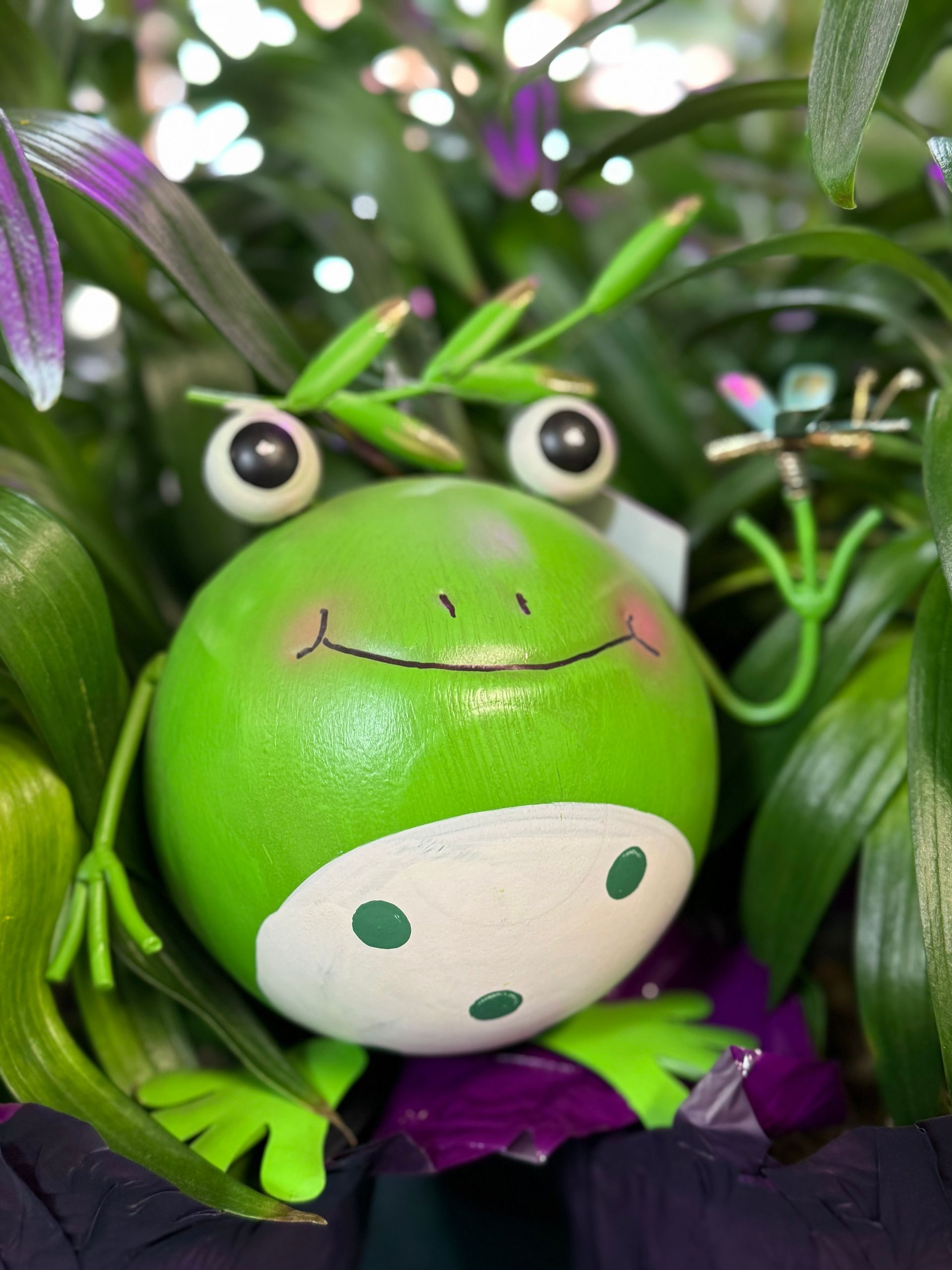 Smiling green frog statue with big eyes, holding a dragonfly. Surrounded by plants.
