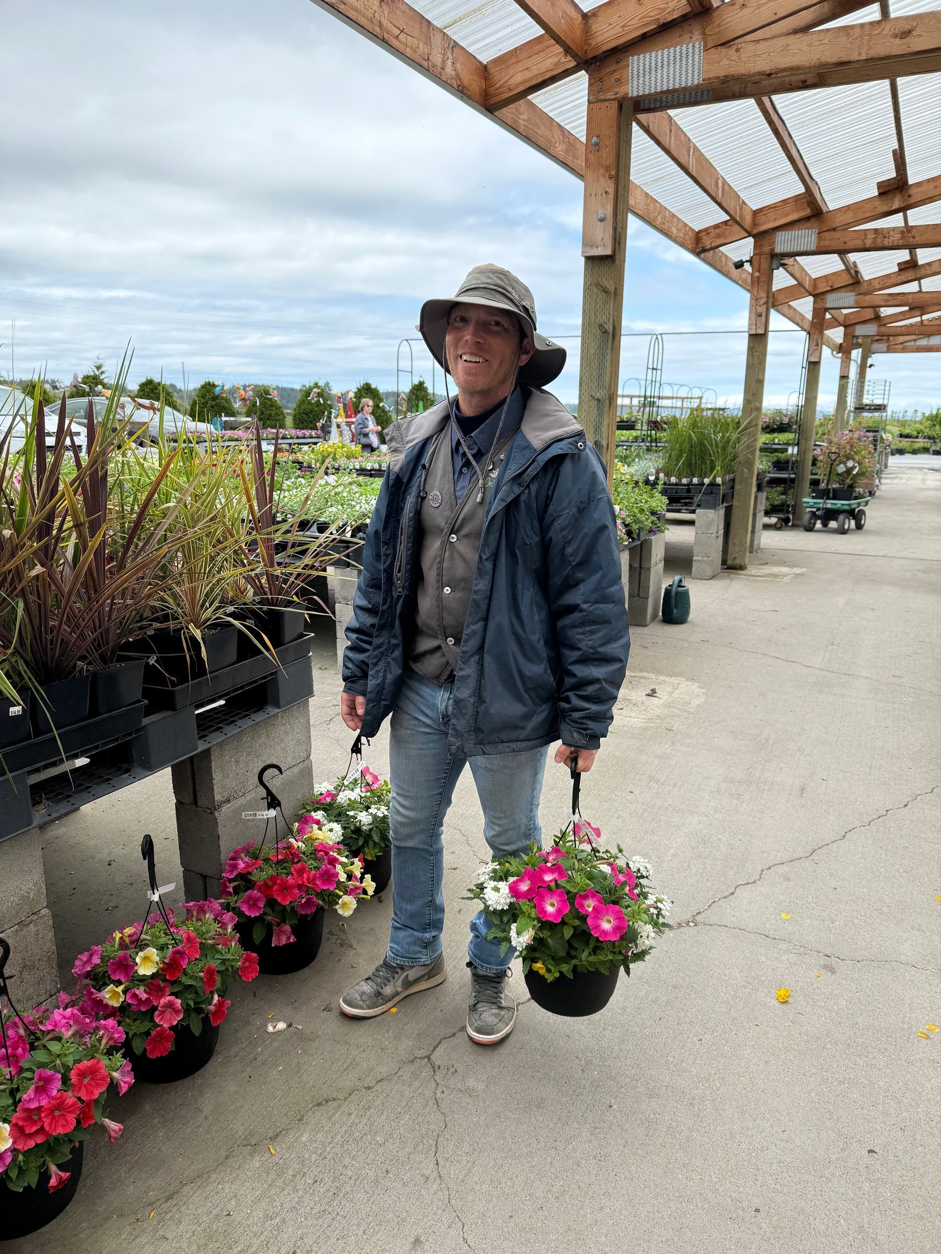 Man in a garden center holding a hanging basket of pink and white flowers.