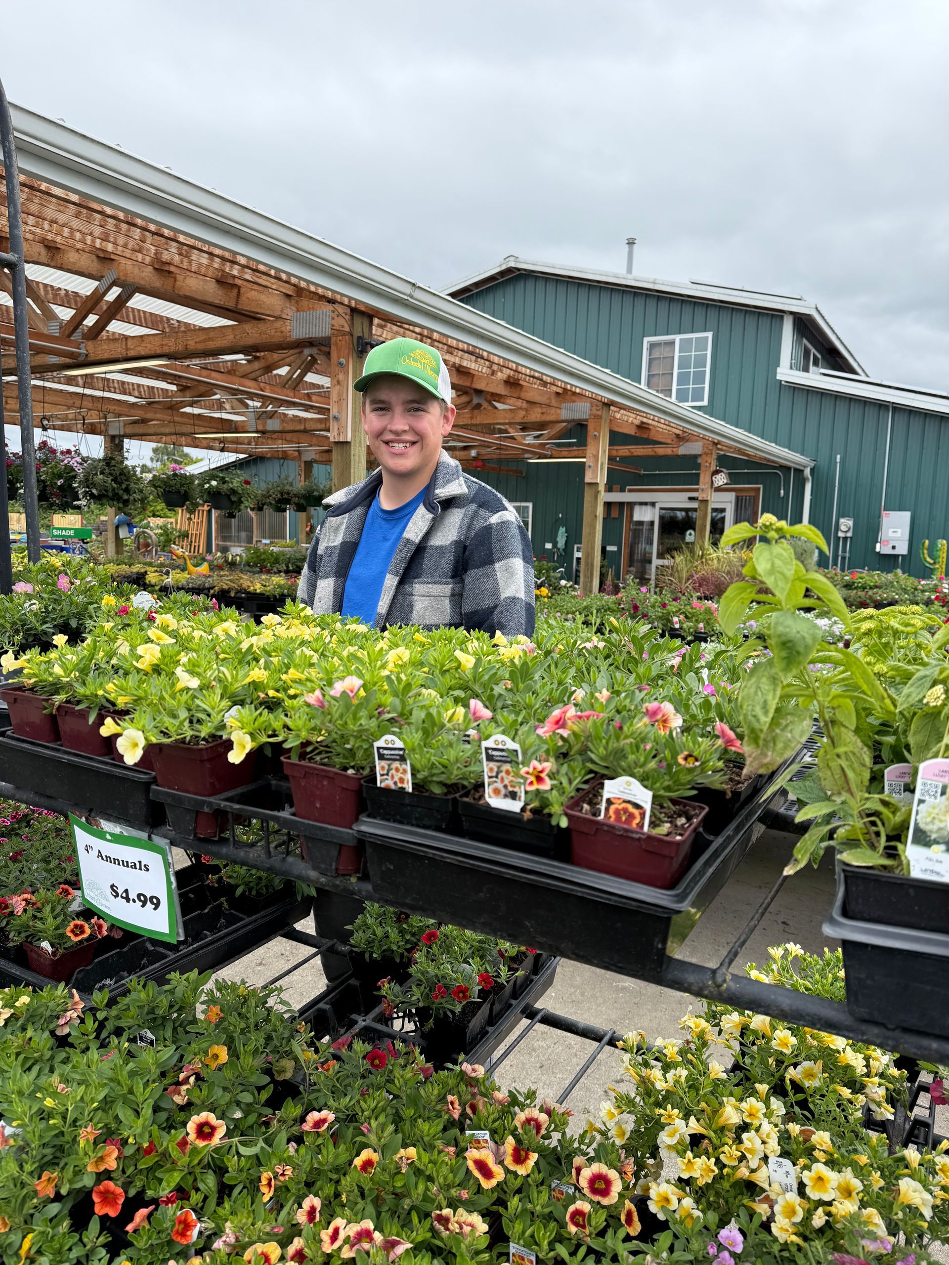 A person smiles, surrounded by flower trays in a garden center. Green hat and plaid shirt.