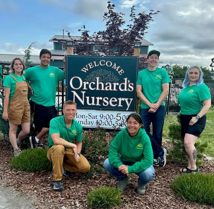 Group of nursery employees wearing green shirts and aprons pose in front of the Orchard's Nursery sign.