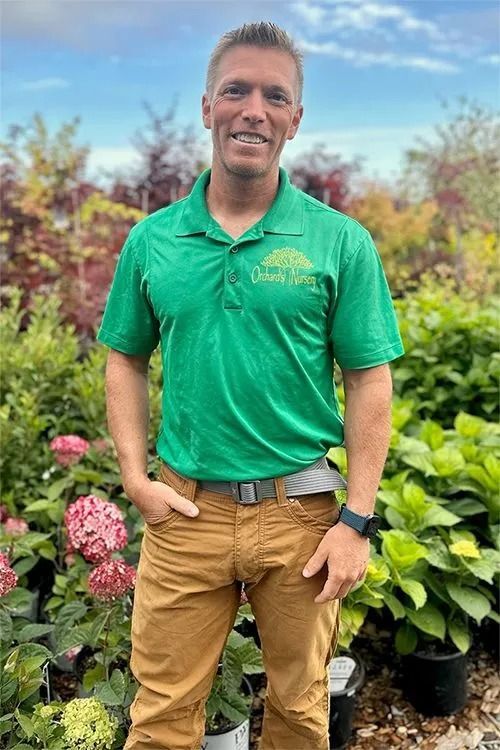 Man in green shirt and tan pants, smiling outdoors among plants.