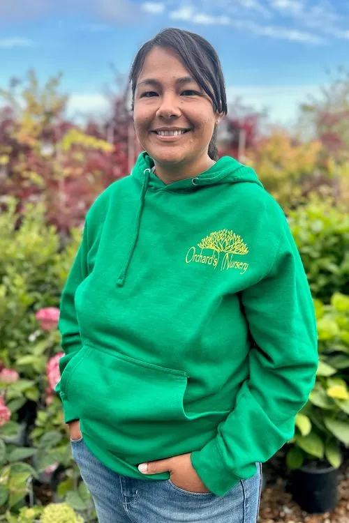 Woman in green hoodie, hands in pockets, smiles in a garden setting.