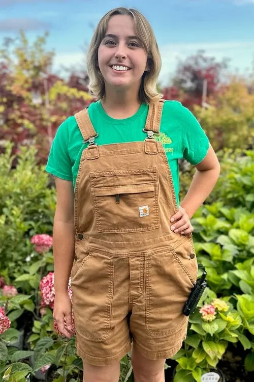 Woman in brown overalls and green shirt smiles outdoors, surrounded by plants.