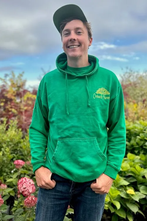 Man in green hoodie and cap, smiling outdoors, hands in pockets.
