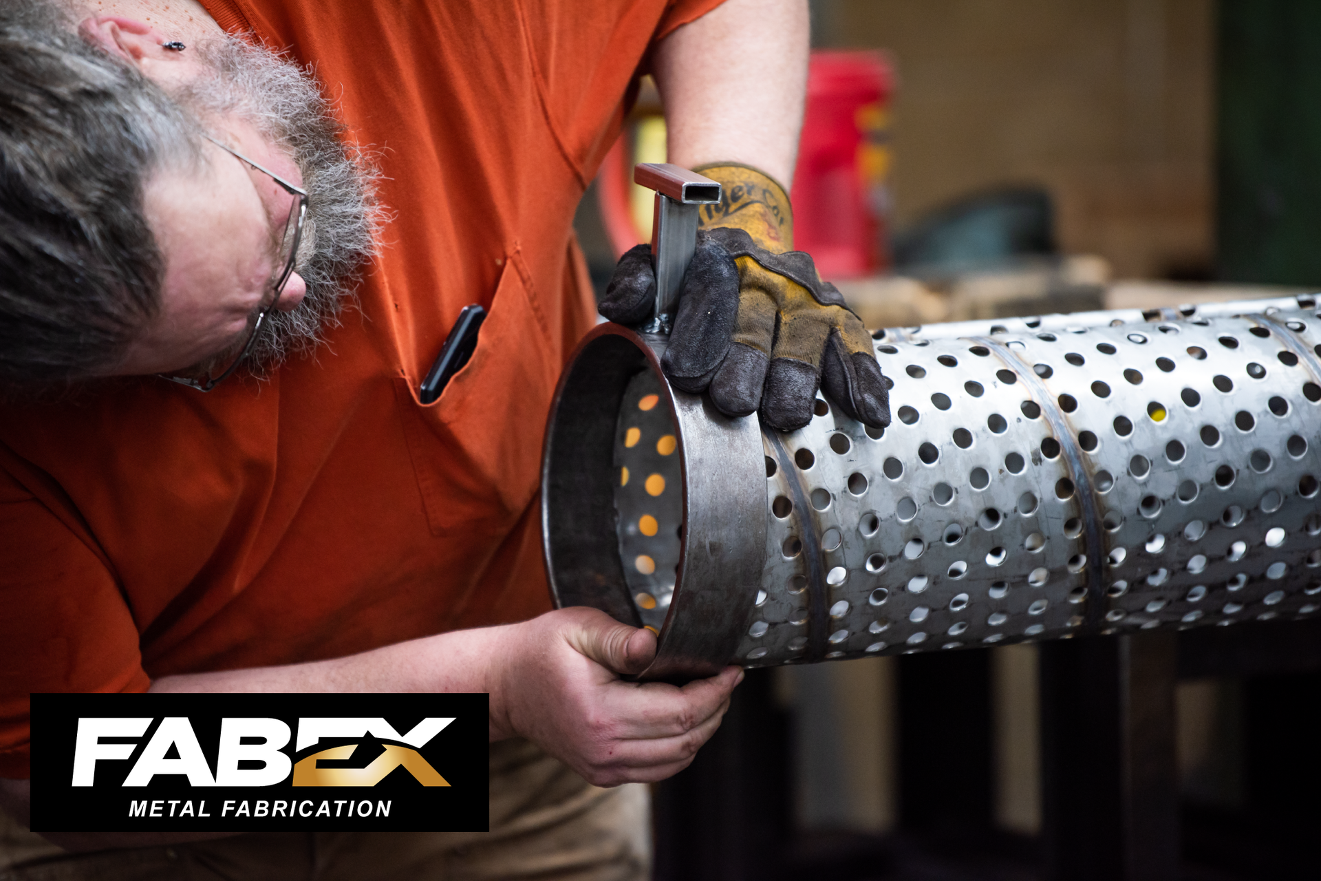 Worker handling a perforated metal cylinder in an industrial workshop, wearing orange shirt and gloves.