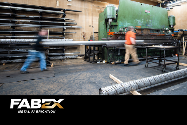 Industrial workshop with workers moving long metal pipes beside a large green machine, FABEX logo visible