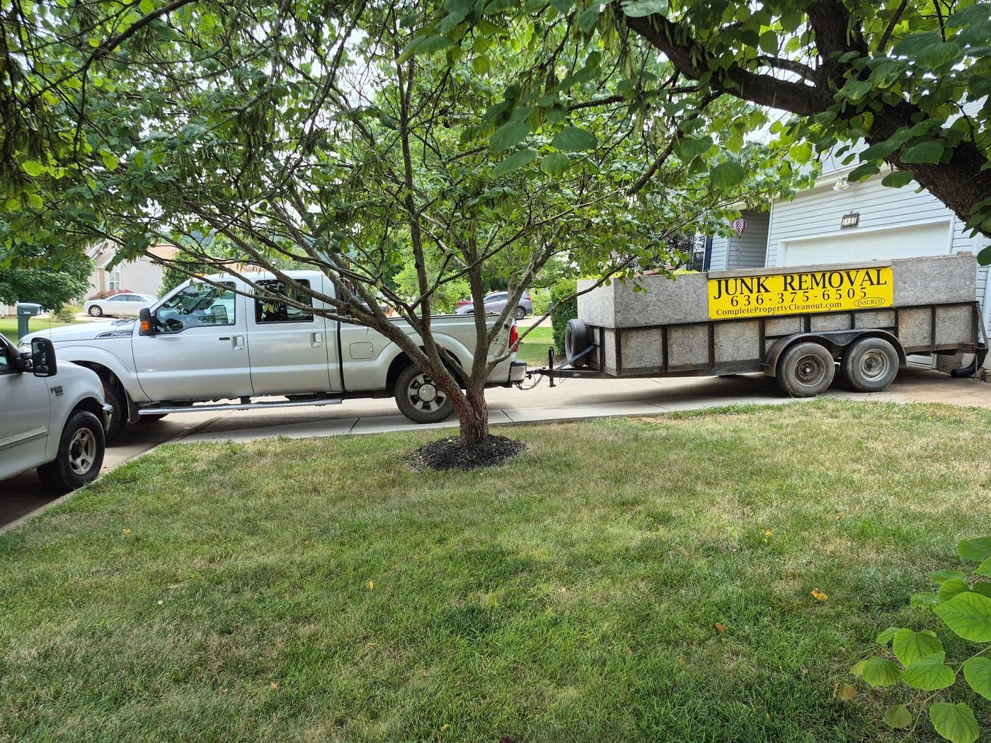 White truck and trailer parked on a lawn, tree in the center. Trailer has a sign for removal services.