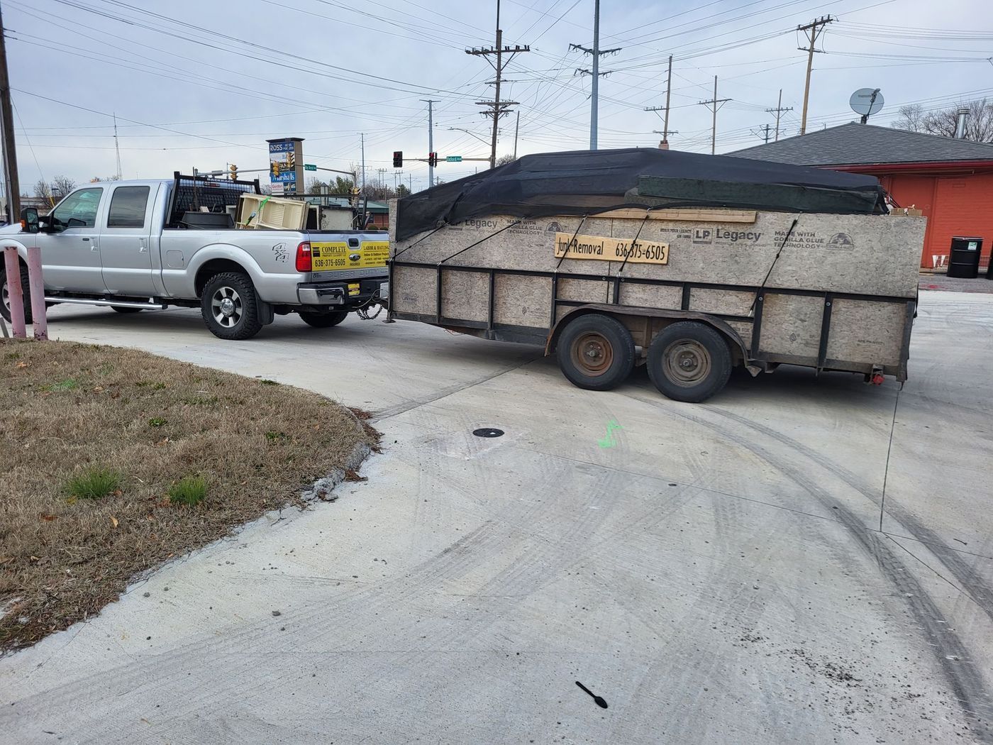 Silver truck towing a trailer covered with black tarp; parked on concrete near a small building with an orange facade.