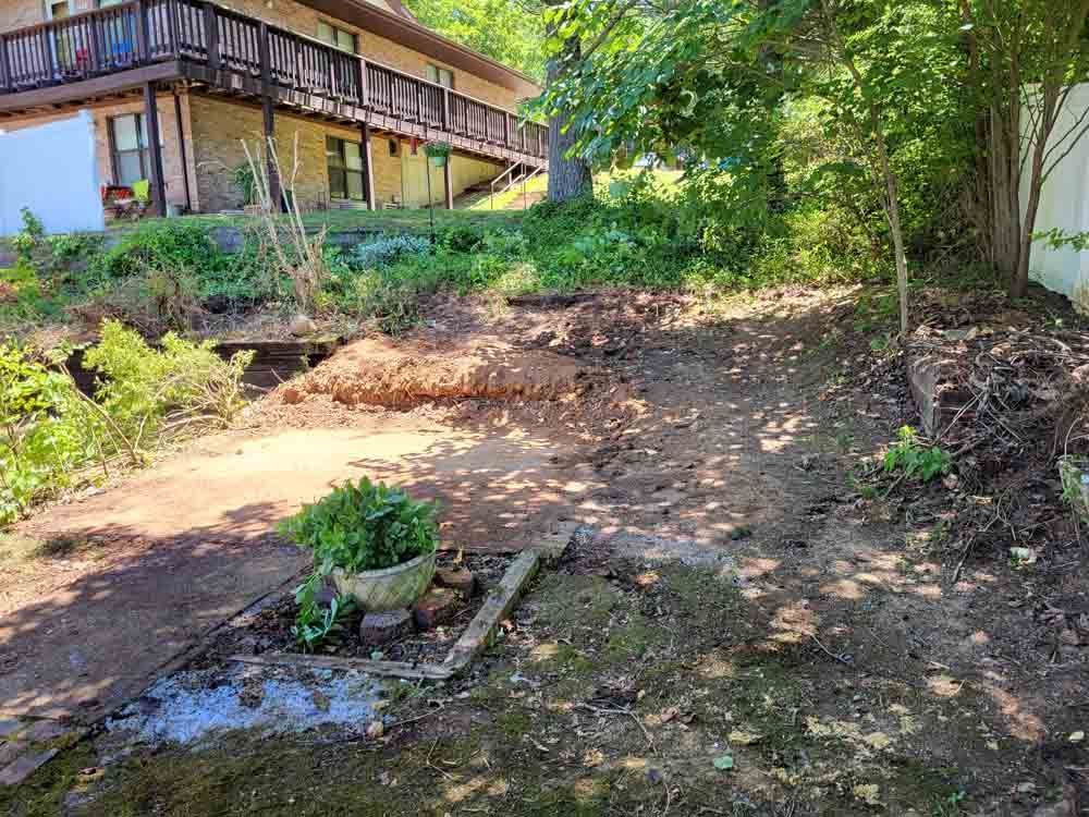 A sloped yard with a building in the background; dirt and greenery. A concrete planter sits in the foreground.