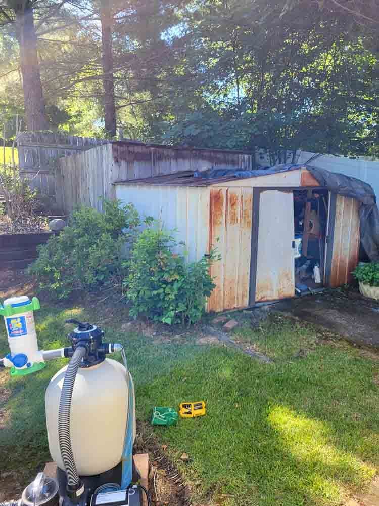 Rusty shed in a backyard with a pool filter on the grass. Trees and a fence are in the background.