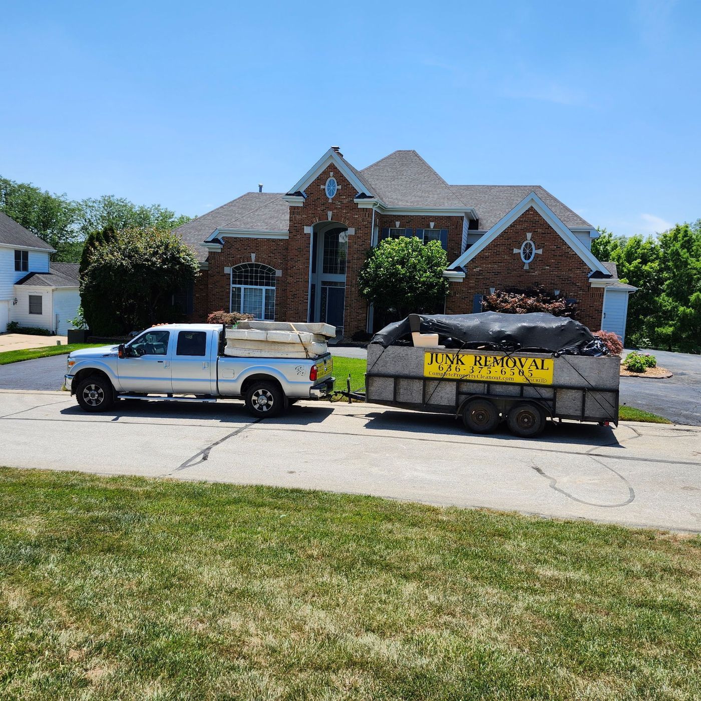 Silver pickup truck towing a trailer filled with debris in front of a large house on a sunny day.