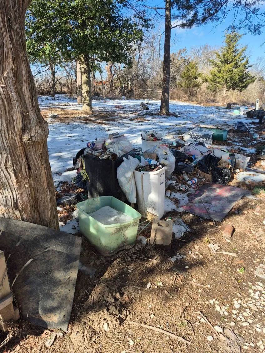 Garbage and debris pile near a tree in a snowy field on a sunny day.