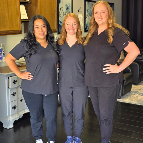 Three people in navy scrubs standing together in a salon, smiling with hands on hips.