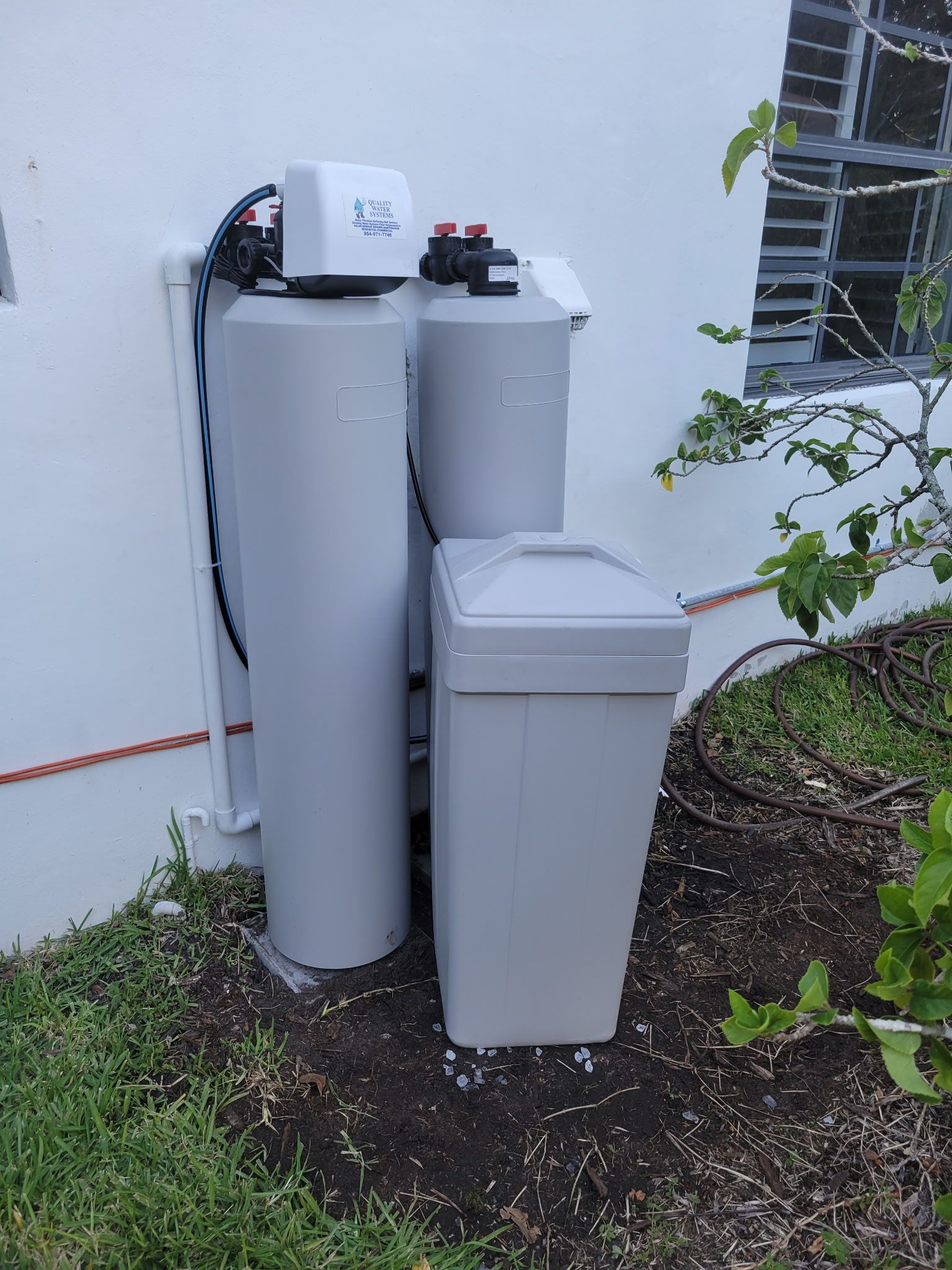 Outdoor utility tanks and bins beside a house wall, with a window and plants nearby.