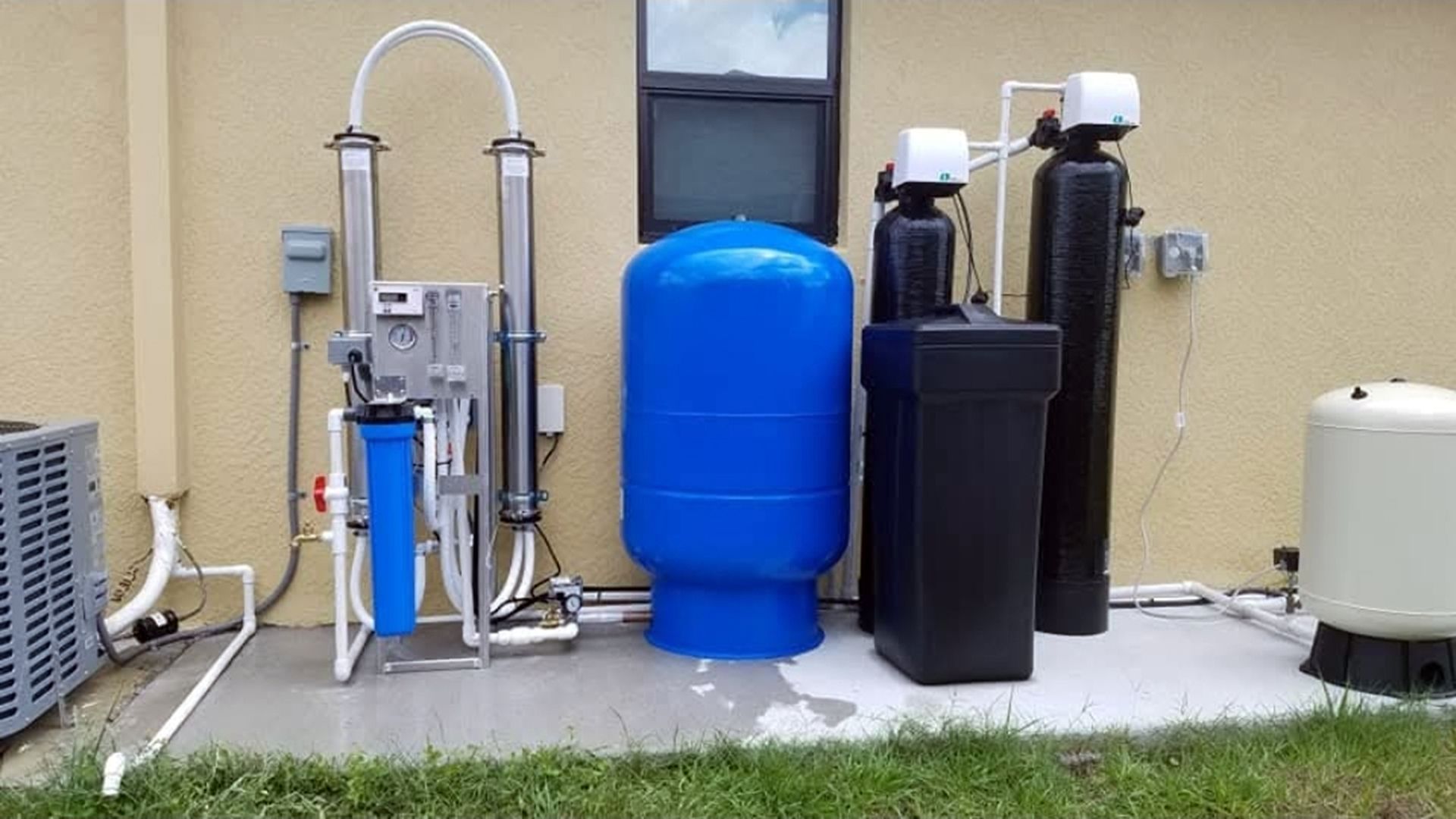 Outdoor water treatment equipment with blue tank and black filter units beside a house wall