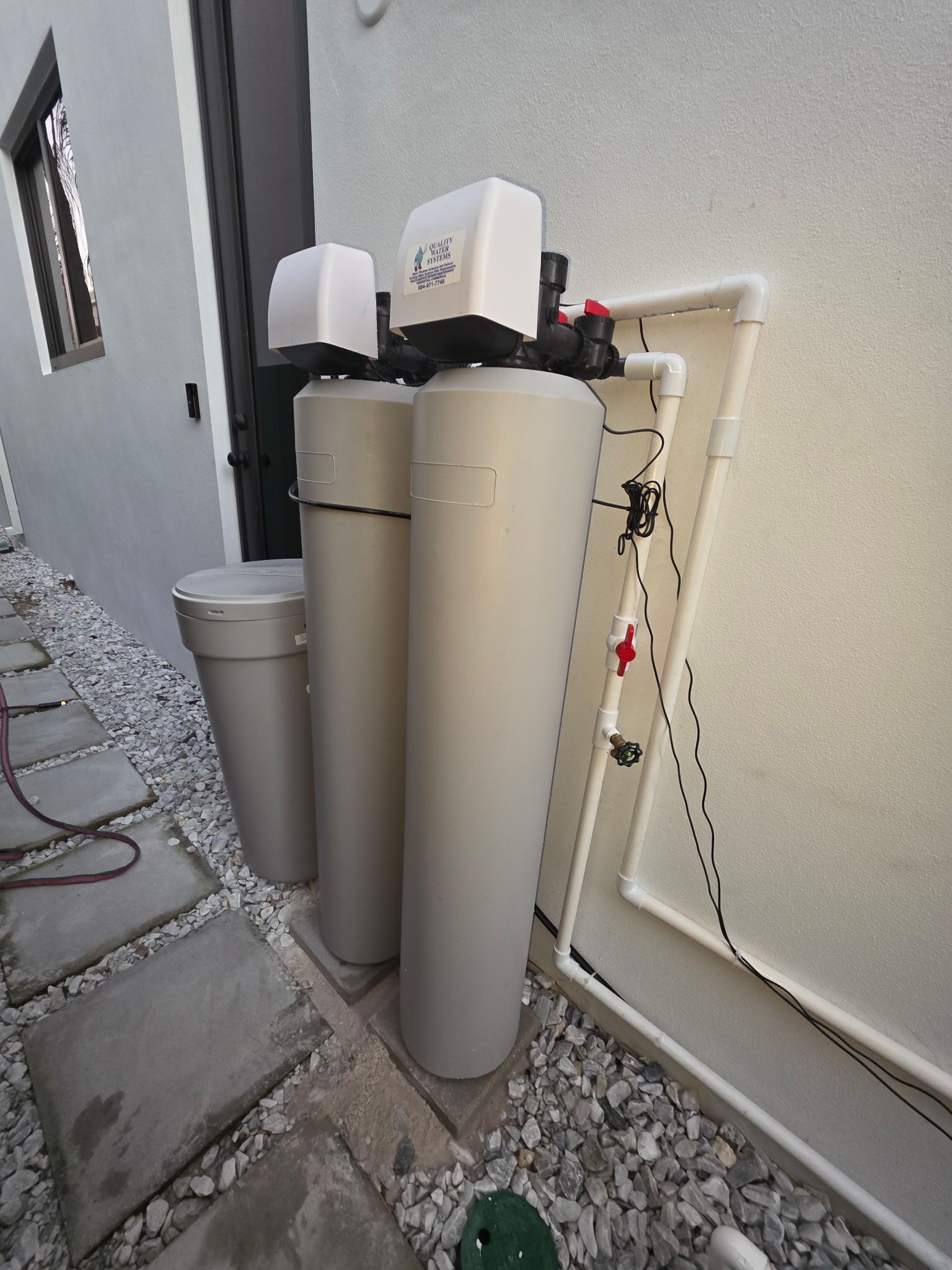 Outdoor water filtration tanks beside a house, with gray and beige cylindrical units and pipes on a gravel strip.
