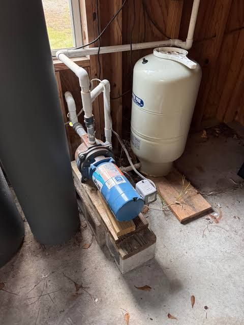 Water pump and pressure tank installed on wooden blocks in a utility corner with exposed pipes.