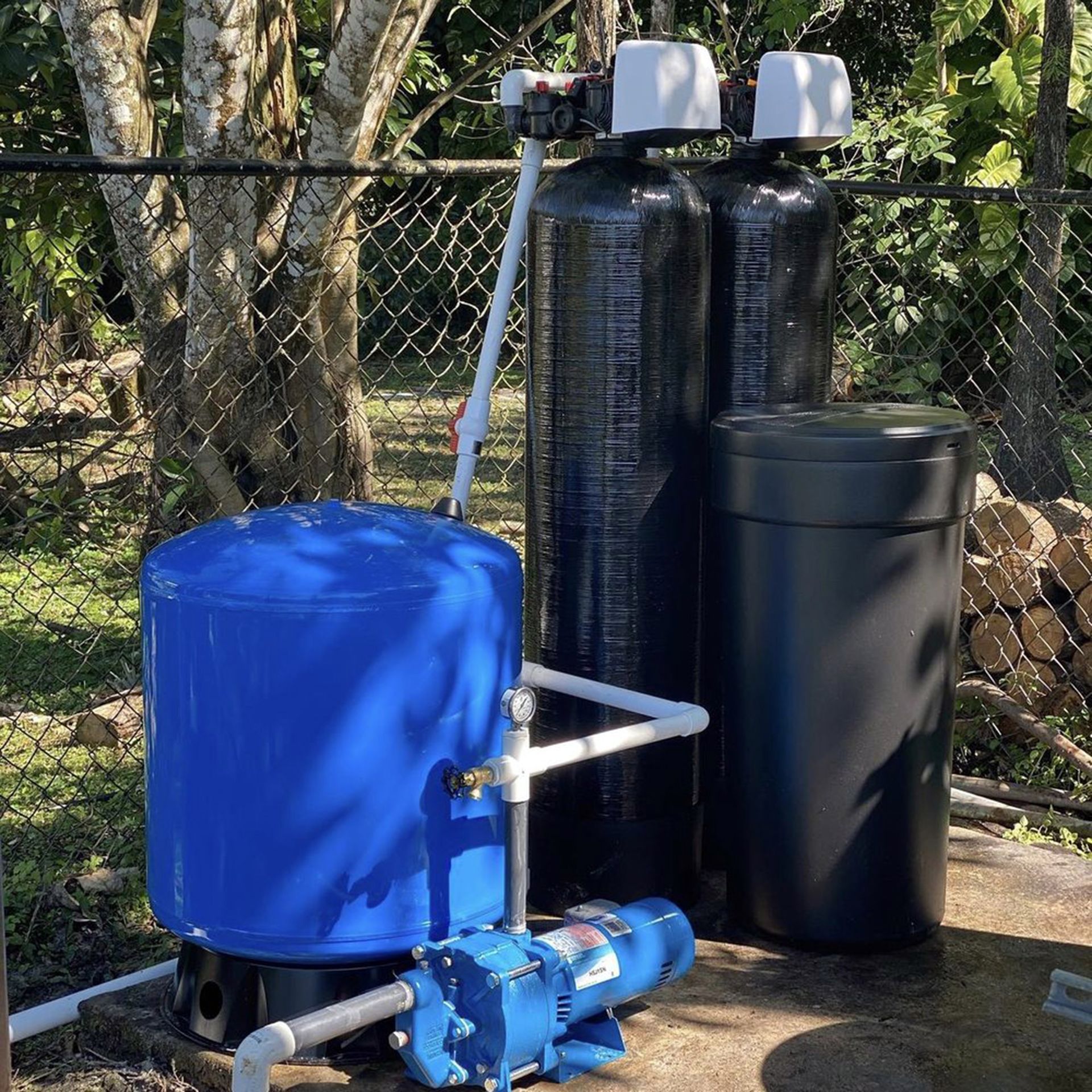 Blue water tank with pump and black storage tanks outdoors by a chain-link fence
