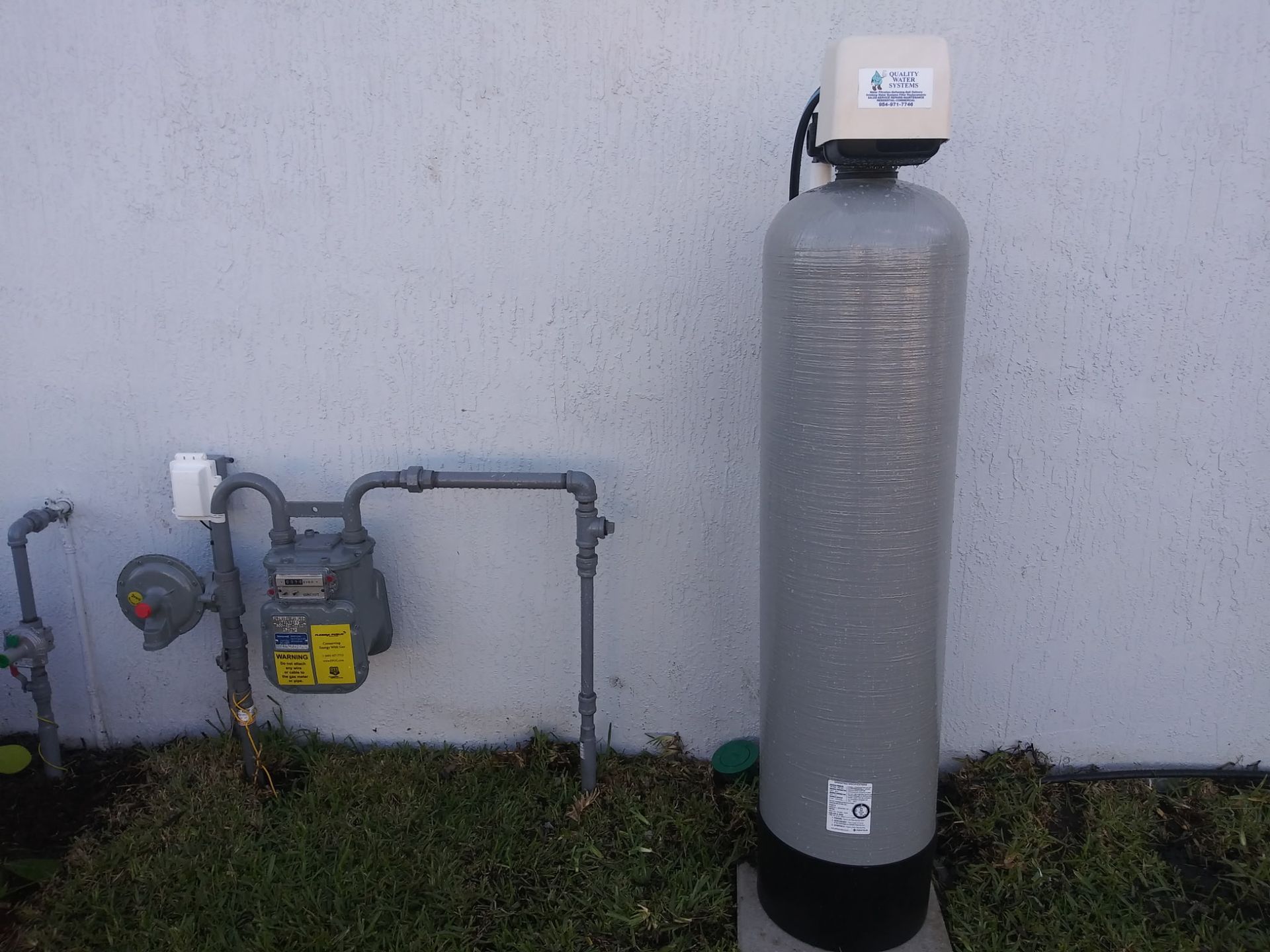 Outdoor gas meter and piping mounted beside a wall, with a tall gray propane tank nearby.