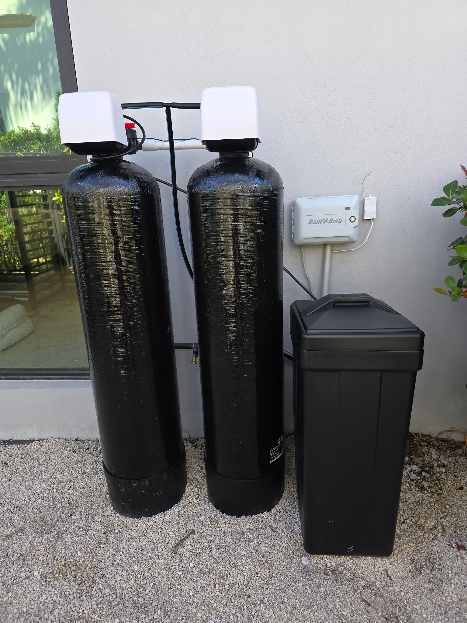Two black water softener tanks and a black brine tank beside a wall outdoors.