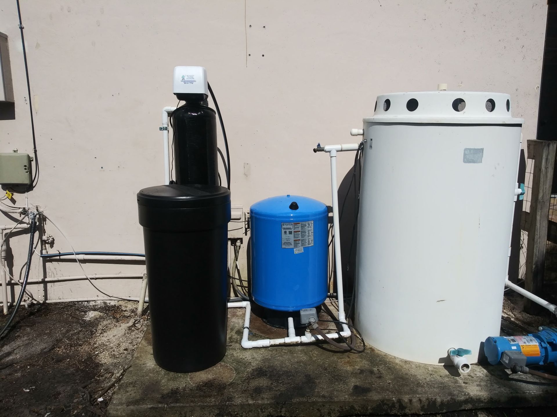 Water treatment equipment: a black tank, blue pressure tank, and white water heater beside a wall.