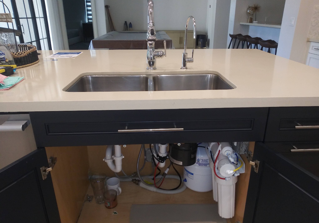 Kitchen island with stainless sink and faucet, dark cabinets, and plumbing visible underneath