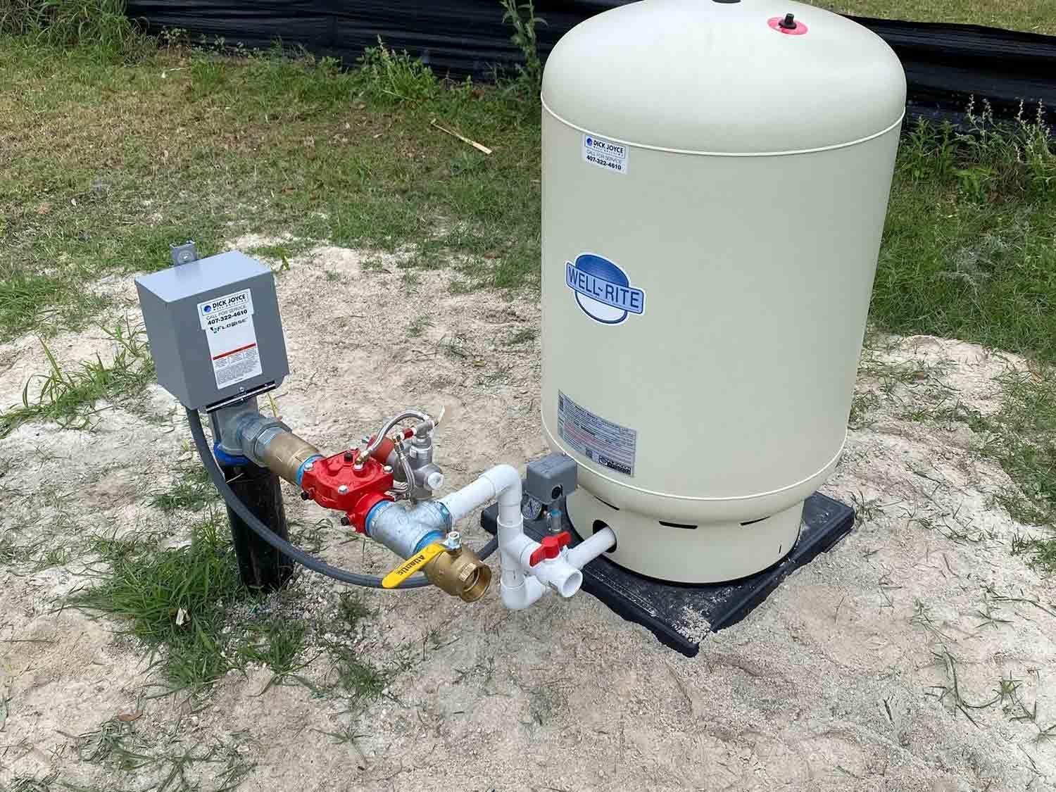 Outdoor well pump and pressure tank setup on gravel, with gray control box and white tank connected by pipes.