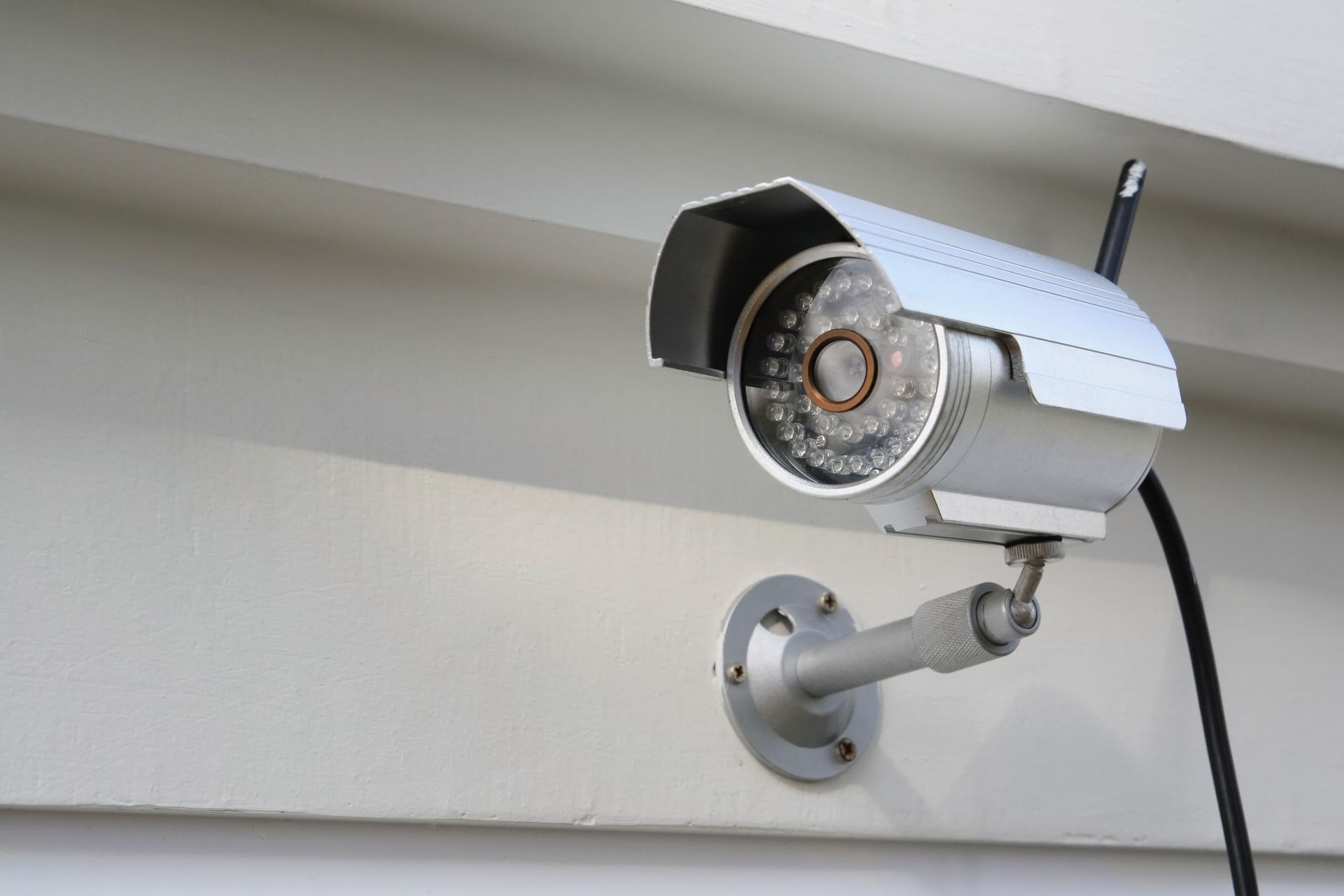A silver outdoor security camera mounted on a white wall, featuring a lens surrounded by small infrared lights.