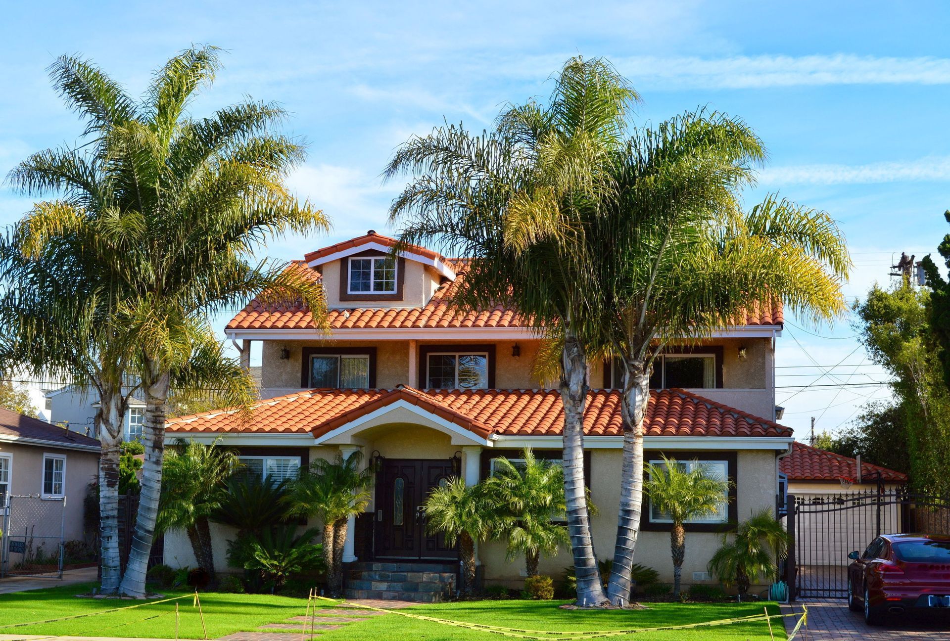 A two-story stucco house with a terracotta tile roof and dormer window, framed by two tall palm trees on a sunny day.