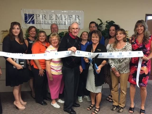 A group of people standing behind a ribbon in front of a Premier Residential Services sign during a ribbon-cutting ceremony.